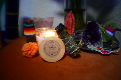 Home altar tabletop with a lit scented candle in a glass jar, tied sage smudge stick, purple amethyst crystal, wooden-lidded jar, orange rose soap and a small plush hummingbird decorative accent