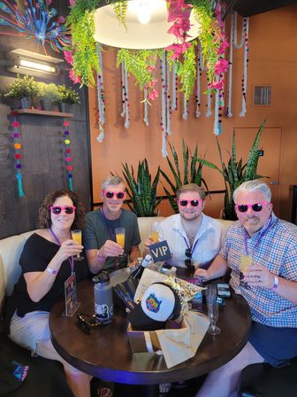 Group of four wearing bright pink sunglasses toasting mimosas at a VIP table in a tropical‑themed indoor lounge with hanging ferns, pink flowers, colorful pom‑pom ropes and snake plants.