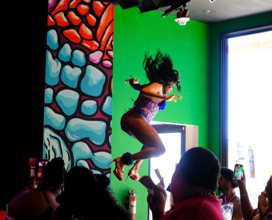Dancer mid-air in a purple cutout costume and platform heels against a bright green wall and colorful mural as an audience films on smartphones