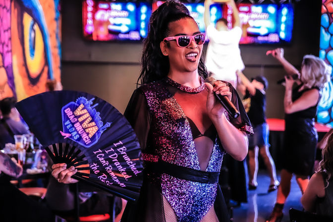 Drag performer in a sparkly purple outfit and pink sunglasses sings into a microphone while holding a fan that says 'Drag Brunch', entertaining a lively crowd under colorful neon lights.