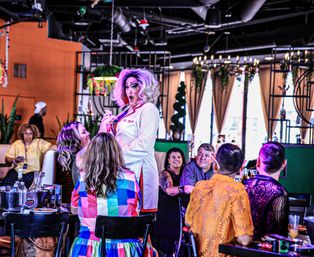 Drag performer in a purple wig and dramatic makeup entertaining a lively restaurant crowd during a colorful drag brunch show, guests laughing and clapping.