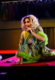 Drag performer with a blonde wig and bold makeup kneeling on a red stage, singing into a microphone in a colorful patterned dress with green feather trim at a lively cabaret show.