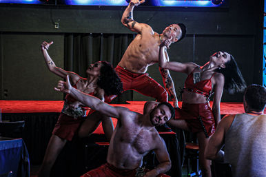 Energetic stage performance with a shirtless male and multiple dancers in red outfits striking dramatic, synchronized poses under colorful LED lighting on a club stage
