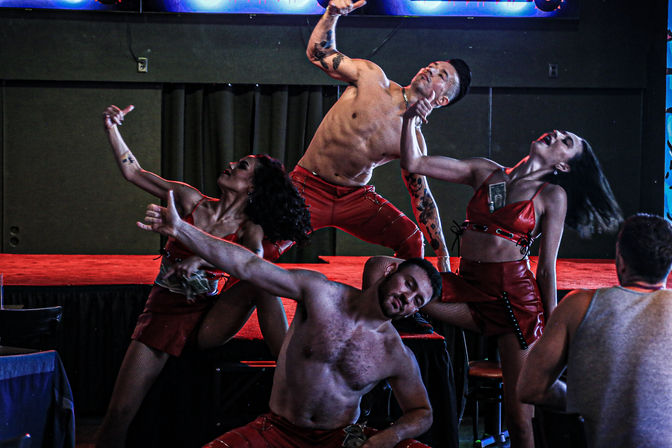 Energetic stage performance with a shirtless male and multiple dancers in red outfits striking dramatic, synchronized poses under colorful LED lighting on a club stage