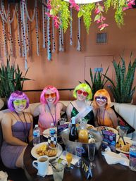 Four friends at a festive indoor brunch wearing colorful wigs (purple, pink, neon green, orange) and sunglasses, seated around a table with plates, mimosas, a bottle in an ice bucket, a VIP sign, hanging plants and rope decorations.
