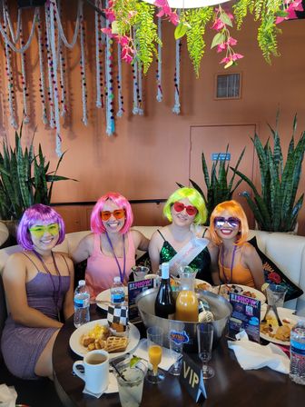 Four friends at a festive indoor brunch wearing colorful wigs (purple, pink, neon green, orange) and sunglasses, seated around a table with plates, mimosas, a bottle in an ice bucket, a VIP sign, hanging plants and rope decorations.