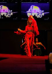 Model on a runway stage wearing a red fringe cowgirl outfit with leopard-print accents, posing in front of large neon palm-sunset visuals on screens during a fashion performance.