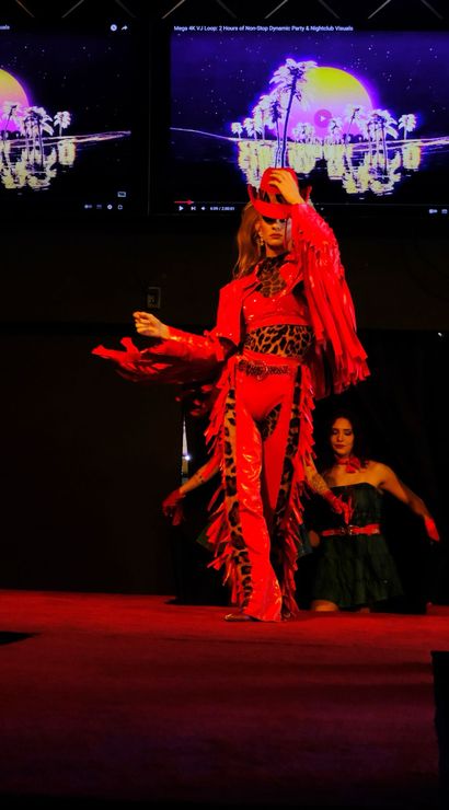 Model on a runway stage wearing a red fringe cowgirl outfit with leopard-print accents, posing in front of large neon palm-sunset visuals on screens during a fashion performance.