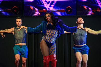 Drag performer in a blue cutout bodysuit and red thigh-high boots leads two backup dancers in mesh tops and blue shorts on a club stage with neon purple LED visuals.