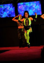 Energetic stage performer in neon green and black lace-up costume and boots dances with a backup dancer in camouflage in front of vivid blue-purple LED visuals on a red runway