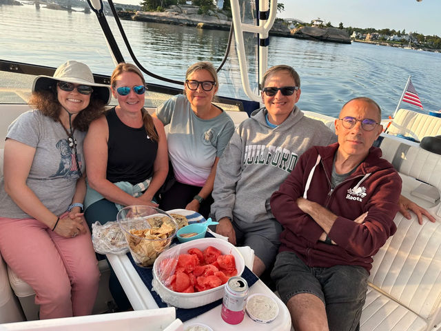 Five adults smiling together on a motorboat in a calm coastal harbor, sharing snacks like watermelon, chips and dip on a small table, rocky shoreline and waterfront homes in the background with an American flag at the stern.