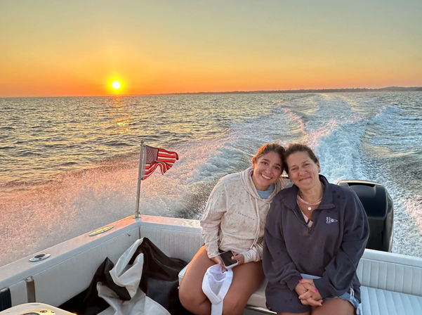 Two people smiling on the stern of a motorboat at sunset, American flag fluttering and the boat wake trailing across the golden ocean under a clear sky.