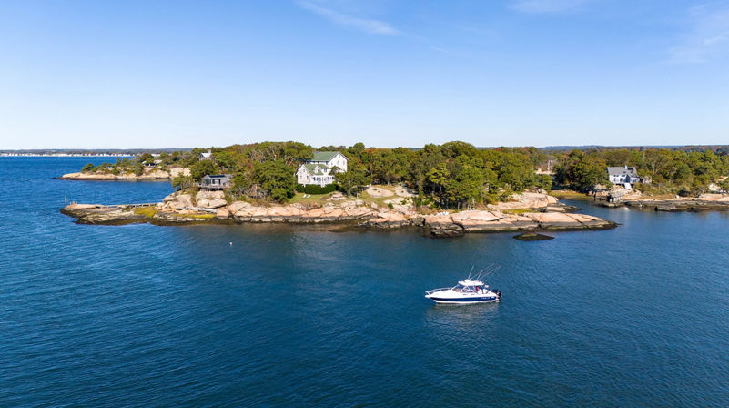 Aerial view of a small tree-covered rocky island with white coastal houses on a granite shore and a motorboat cruising on calm blue water under a clear sky.