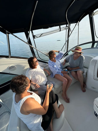 Four women lounging in a boat’s shaded cockpit, sipping drinks and chatting with calm open sea and a small island on the horizon