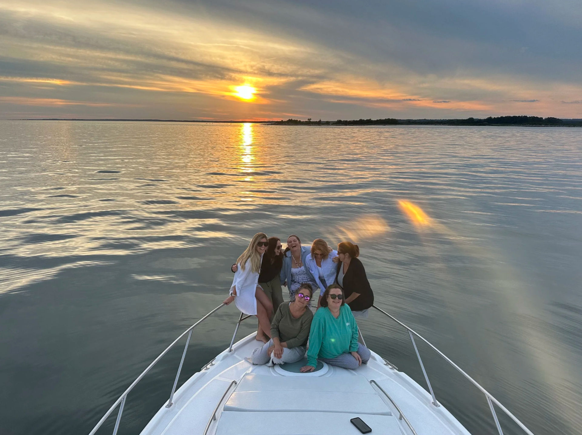 Group of friends relaxing on a boat bow at sunset, golden sun and colorful sky reflecting across calm water with a distant shoreline — serene sunset boat cruise scene.