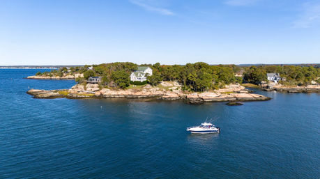 Aerial view of a rocky coastal island with trees and a white waterfront house, a motorboat anchored in calm blue sea under a sunny clear sky.