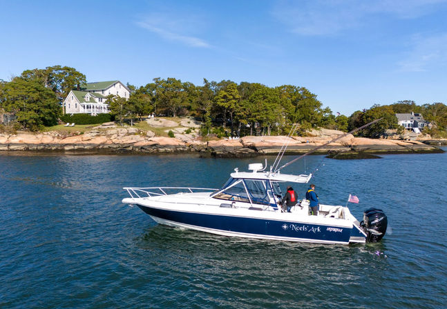 Blue-and-white motorboat with two anglers fishing in calm nearshore waters, passing a rocky tree-lined shoreline with coastal homes under a clear blue sky.