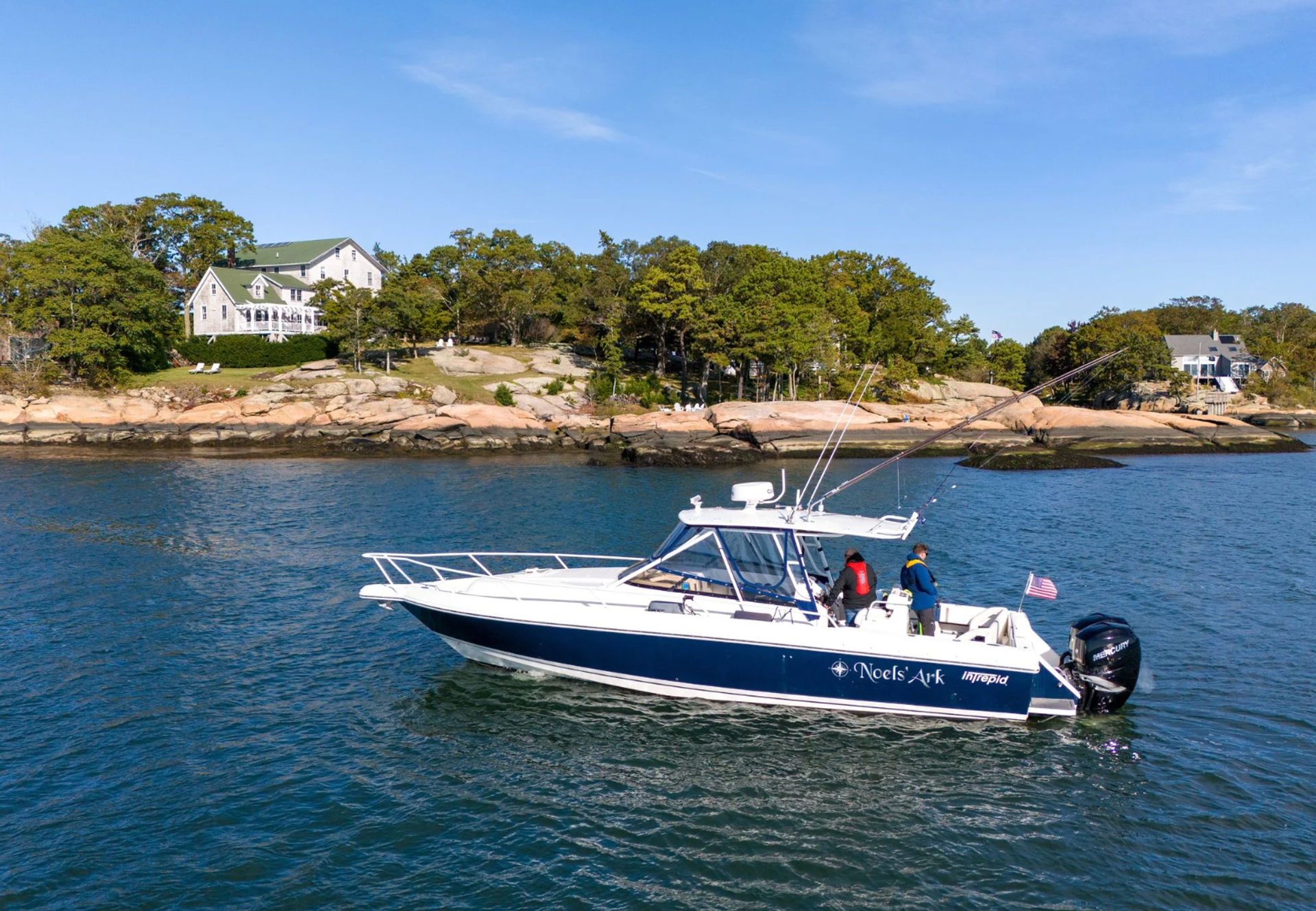 Blue-and-white powerboat with fishing rods and a small American flag cruising in calm coastal waters past a rocky, tree-lined shoreline and seaside homes under a clear blue sky.