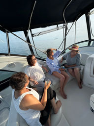 Group of four women relaxing with drinks in a covered motorboat cockpit on calm open water, clear sky and a small island on the horizon.