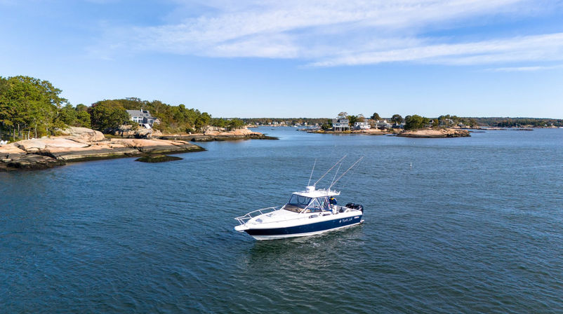 White-and-blue motorboat cruising through a calm coastal inlet, passing rocky granite shorelines and waterfront homes under a bright blue sky