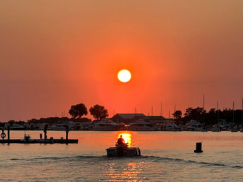 Small motorboat with silhouetted boater heading toward a vivid orange sunset over a marina, sun reflecting on calm harbor waters with yachts and shoreline trees.