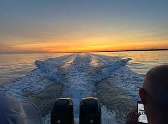 Golden sunset over calm water with a boat's twin outboard engines leaving a V-shaped wake toward the distant horizon