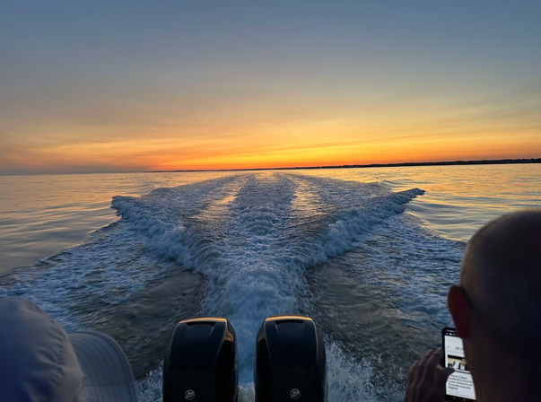 Golden sunset over calm water with a boat's twin outboard engines leaving a V-shaped wake toward the distant horizon
