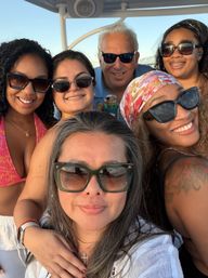 Sunset boat selfie of six friends wearing sunglasses and summer outfits, smiling on the water with a bridge in the background.