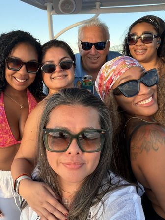 Sunset boat selfie of six friends wearing sunglasses and summer outfits, smiling on the water with a bridge in the background.