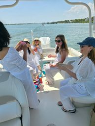 Friends in white outfits laughing and sharing snacks on a sunny summer boat ride in a calm coastal bay with docks and shoreline visible