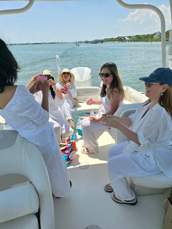 Friends in white outfits laughing and sharing snacks on a sunny summer boat ride in a calm coastal bay with docks and shoreline visible