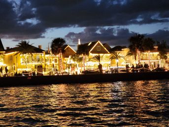 Festive waterfront at night: palm-lined promenade and homes draped in warm holiday string lights, silhouetted strollers and sparkling reflections on rippling water beneath a moody cloudy sky.