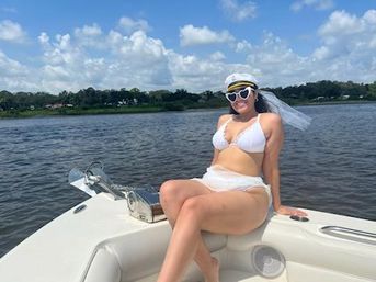 Woman in white bikini wearing a captain's hat with veil and heart-shaped sunglasses relaxing on the bow of a boat on a sunny lake with tree-lined shoreline and puffy clouds