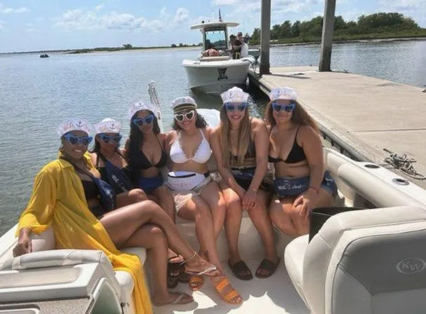 Six friends in swimwear wearing matching sailor hats and sunglasses posing cheerfully on a leisure boat docked at a sunny coastal inlet by a wooden dock