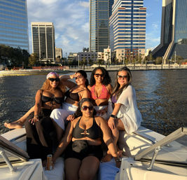 Five friends in swimsuits lounging on a boat near an urban waterfront at golden hour, with modern downtown skyscrapers and calm river in the background.