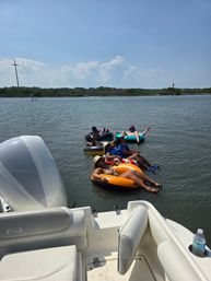 Group of people lounging on colorful inner tubes tied to a boat, floating in a calm coastal inlet near marshy shoreline under a bright blue summer sky with a distant cross-shaped marker