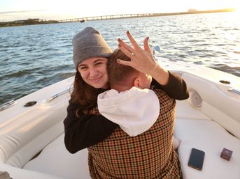 Smiling woman in a beanie hugging her partner on a boat at sunset, proudly showing an engagement ring with water and a distant bridge in the background.