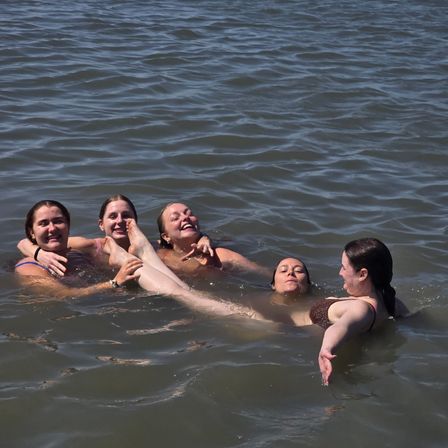Five friends in swimsuits laughing and floating in a lake, supporting one friend horizontally with her legs raised for a playful summer swim.