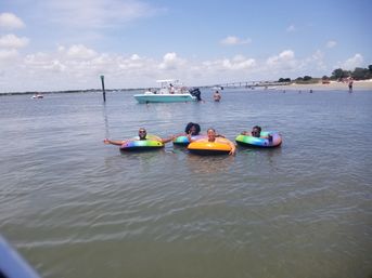 Four people relaxing on colorful inflatable tubes in a calm coastal bay with a nearby anchored boat, distant bridge and sandy beach under a sunny sky