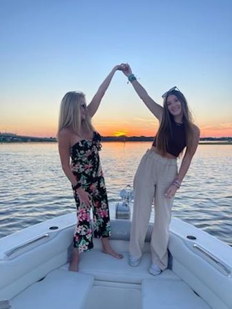 Two friends on a boat at sunset on a calm lake, holding hands overhead and smiling during a summer sunset cruise.