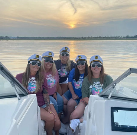 Five friends in captain hats and sunglasses on a boat at sunset, wearing colorful "50th Birthday Trip" shirts with calm water reflecting the sun.