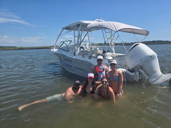 Smiling group of six people and a child wading in shallow coastal waters next to a white recreational motorboat with a canopy under a bright blue sky — summer boating outing on a calm bay.