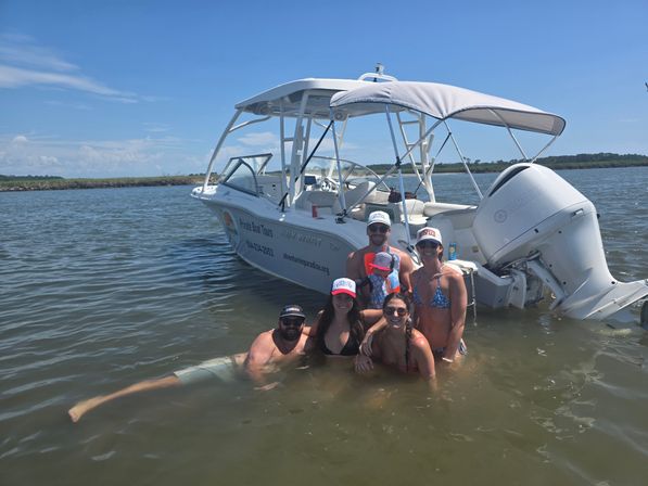 Smiling group of six people and a child wading in shallow coastal waters next to a white recreational motorboat with a canopy under a bright blue sky — summer boating outing on a calm bay.