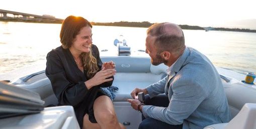 Romantic boat proposal at sunset — a man presents an open ring box to a surprised, smiling woman aboard a small motorboat with calm water and a bridge in the background.