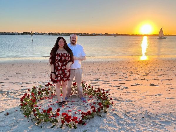 Romantic couple standing inside a heart-shaped ring of red roses on a sandy beach at golden sunset, calm water and a sailboat reflecting the sun.