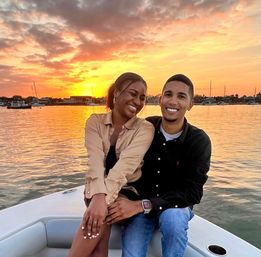 Smiling couple on a boat at sunset, golden sky reflecting on calm harbor waters with sailboats and waterfront houses in the background.