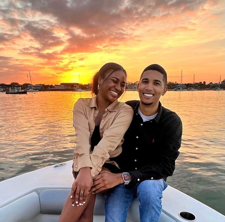 Smiling couple on a boat at sunset, golden sky reflecting on calm harbor waters with sailboats and waterfront houses in the background.