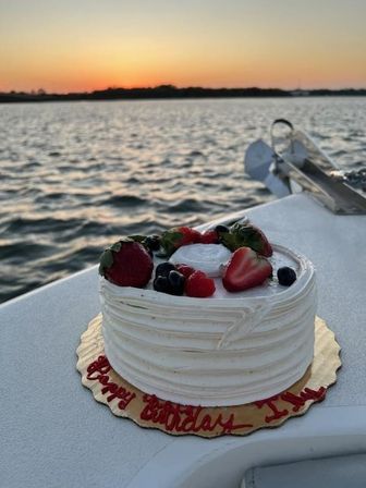 White frosted birthday cake topped with strawberries, blueberries and raspberries, resting on a boat deck with a golden sunset over rippling water in the background.