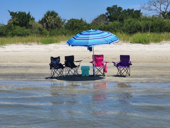 Empty colorful folding beach chairs (two navy, one pink, one purple) under a blue striped umbrella with a teal cooler on a sandy beach shoreline, tidal water reflecting the setup and coastal dunes with green vegetation in the background.