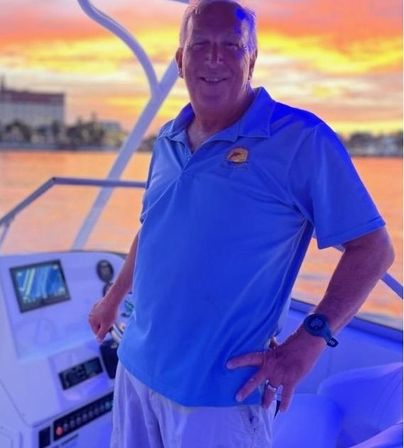 Smiling man at a boat helm in a blue polo, hands on the console with navigation display, vibrant orange-pink sunset over a coastal marina and waterfront skyline